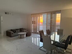 Ein modernes Wohnzimmer mit einer Couch, einem runden Couchtisch und einem Glastisch mit Stühlen. Große Fenster bieten Blick auf die Skyline bei Sonnenuntergang.
