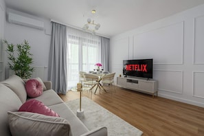 Ein modernes Wohnzimmer mit einer Couch, einem Esstisch mit Stühlen, einem Fernseher, der 'NETFLIX' anzeigt, und einem großen Fenster mit Vorhängen.
