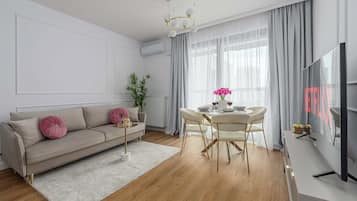 Ein modernes Wohnzimmer mit Sofa, Esstisch, Stühlen, Fernseher und einem großen Fenster mit Vorhängen.