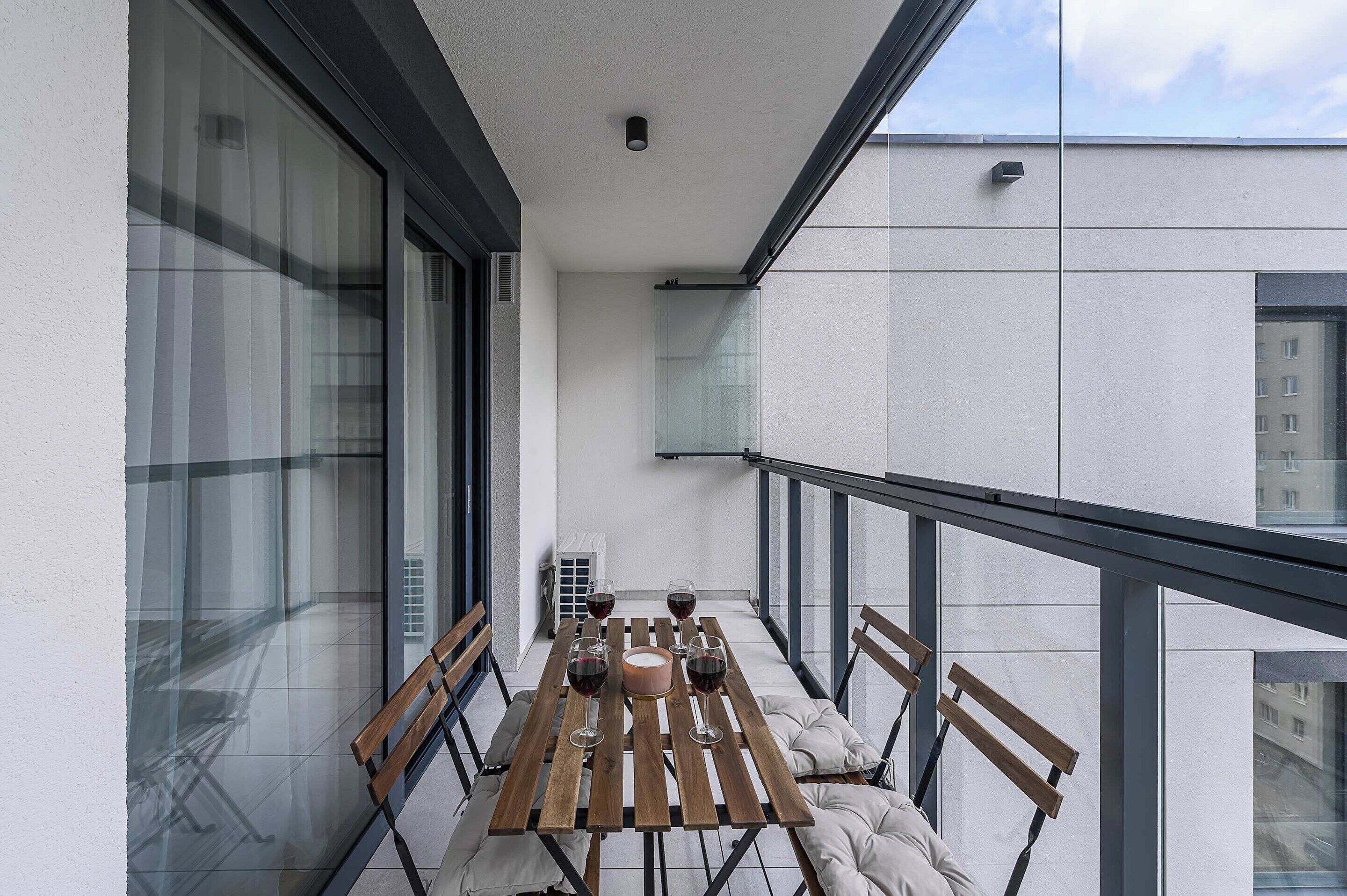 Uma varanda com mesa e cadeiras de madeira, janelas amplas e vista para a paisagem urbana.