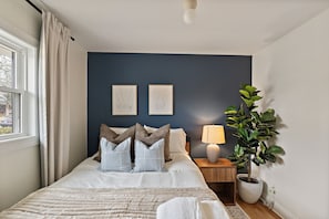 A bedroom with a bed, two wall-mounted pictures, a bedside table with a lamp, and a potted plant. - Luxury Stay with Hot Tub, Fire Pit and Games (Charlotte)