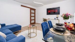 A modern living room with a blue sofa set, a glass coffee table, and a dining area with a TV displaying 'NETFLIX' in red letters. - City Apartment in Warsaw (Warsaw)