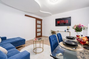 Ein modernes Wohnzimmer mit einem blauen Sofagarnitur, einem Glastisch und einem Essbereich, auf dem Fernseher 'NETFLIX' in roten Buchstaben angezeigt wird.