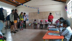 Un aula con estudiantes sentados en pupitres, una profesora al frente y carteles educativos en las paredes.