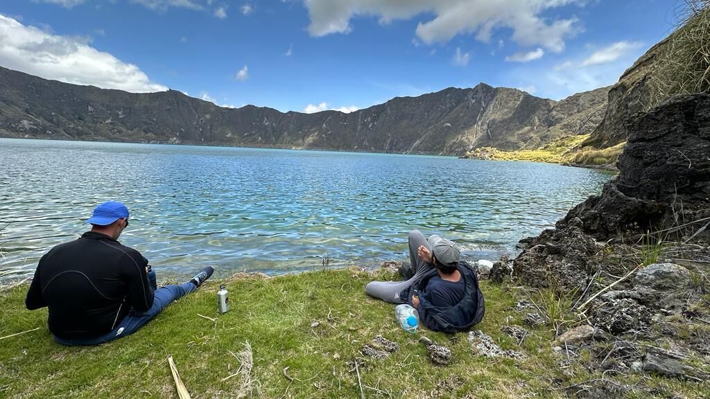 Due persone sedute in riva a un lago, con montagne sullo sfondo.