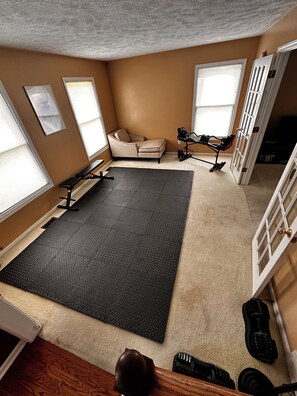 A room with a large exercise mat, a bench, and a chair. There are windows with blinds, a door, and a staircase.