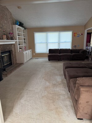 A living room with a fireplace, a brown sofa set, a white shelving unit, and a large window with blinds. - Fountain House- Spacious Okemos home in nature with comforts near MSU (Okemos)