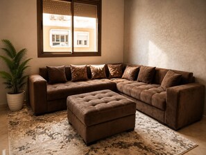 Ein braunes Ecksofa mit passendem Hocker, einer Zimmerpflanze und einem gemusterten Teppich in einem Raum mit einem großen Fenster.