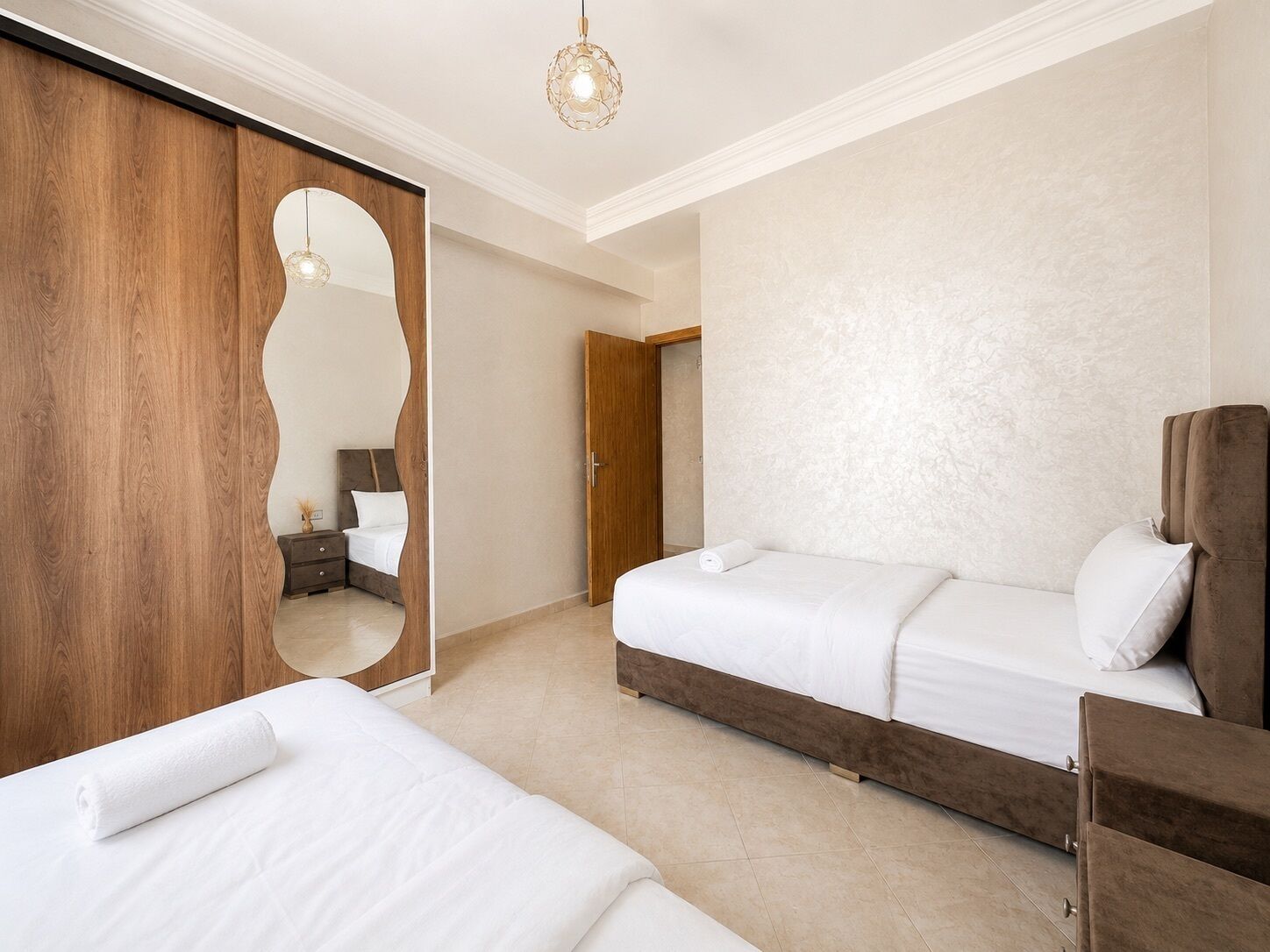 A hotel room with two beds, a wooden wardrobe, a mirror, and a hanging light fixture.