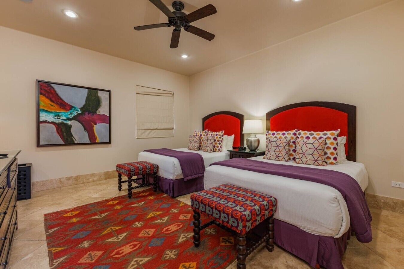 A hotel room with two beds, a ceiling fan, a colorful abstract painting, and a patterned rug.