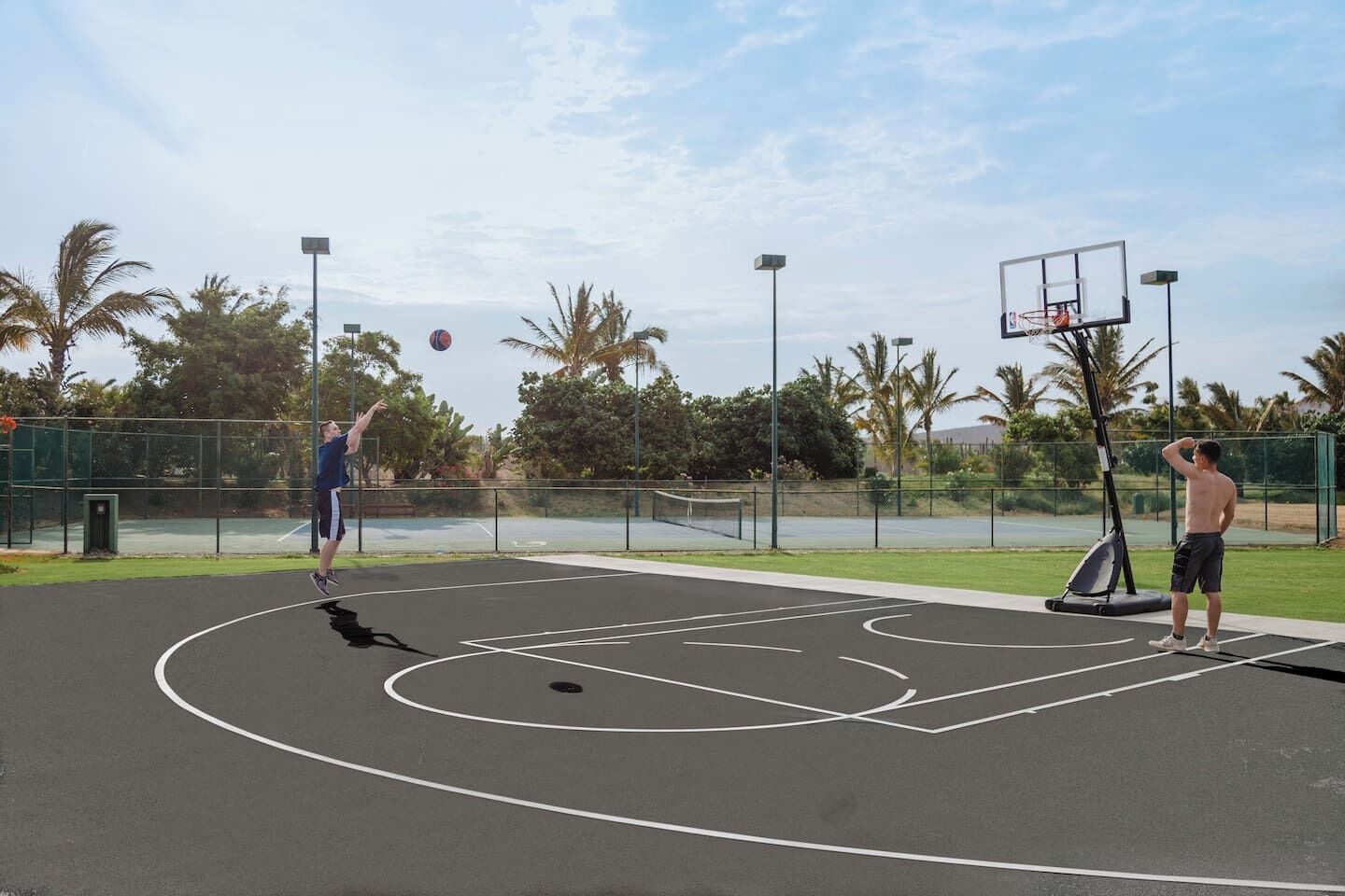 An outdoor basketball court with a hoop, a net, and a basketball in mid-air.