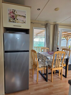 A compact kitchen with a stainless steel refrigerator, wooden dining table with chairs, and a window with curtains. - 3-bedroom mobile home in pleasant Dolus-d'Oléron with AC and 4 x bikes (Dolus-d'Oléron)