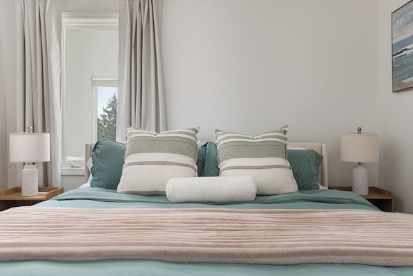 A bedroom with a bed, pillows, a nightstand, a lamp, and a window with curtains.