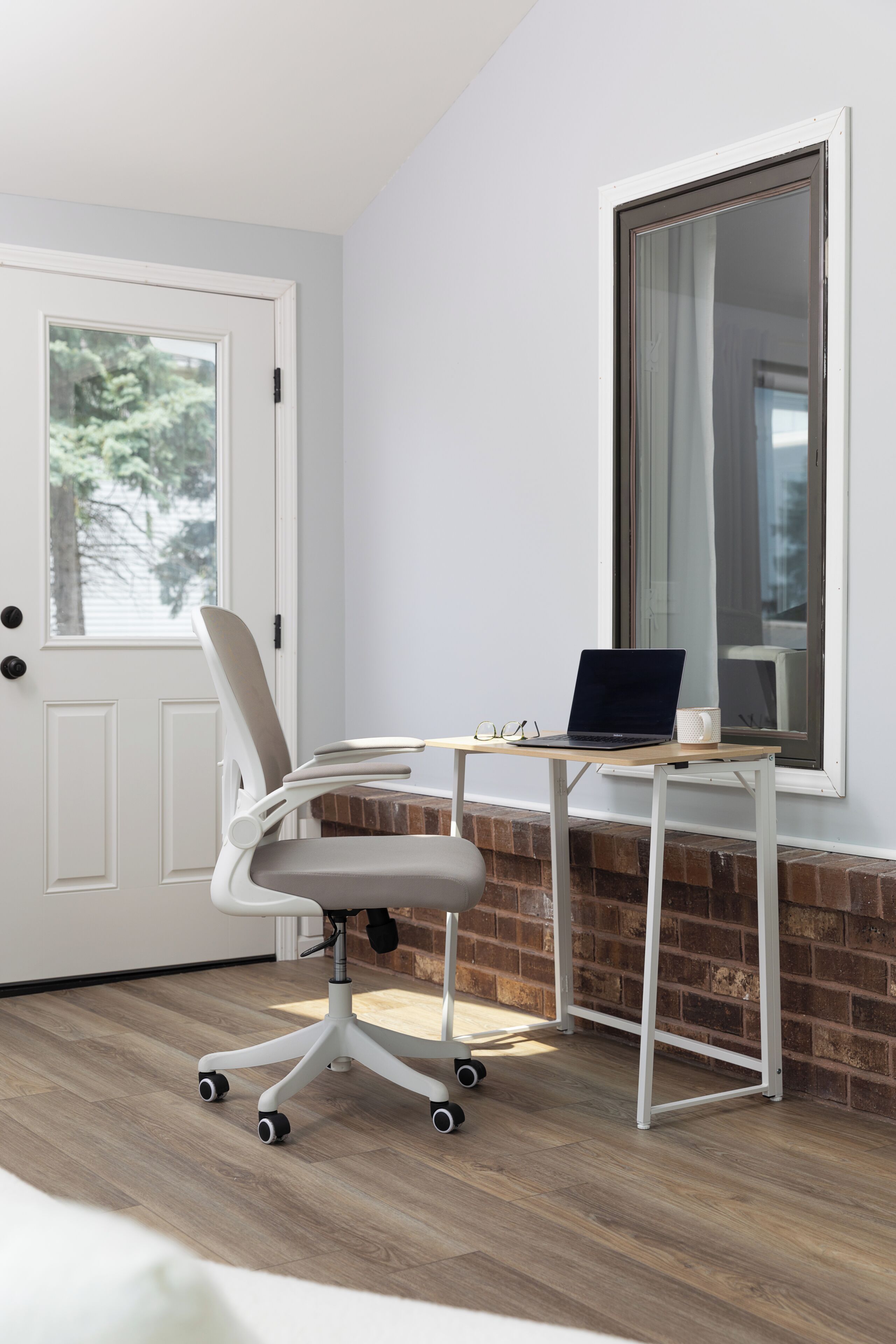 A modern office chair with wheels, a laptop on a desk, and a window with a view of trees.