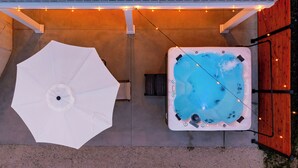Un bain à remous avec un intérieur bleu, un parasol blanc et des guirlandes lumineuses sur une terrasse en béton.