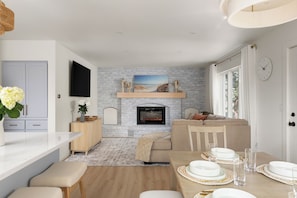 A modern living room with a fireplace, a dining area with a table set for four, and a kitchen with a flat-screen TV.