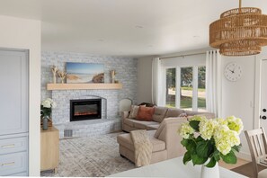 A living room with a fireplace, a sofa, a dining table with a vase of flowers, and a view of the outdoors through large windows.