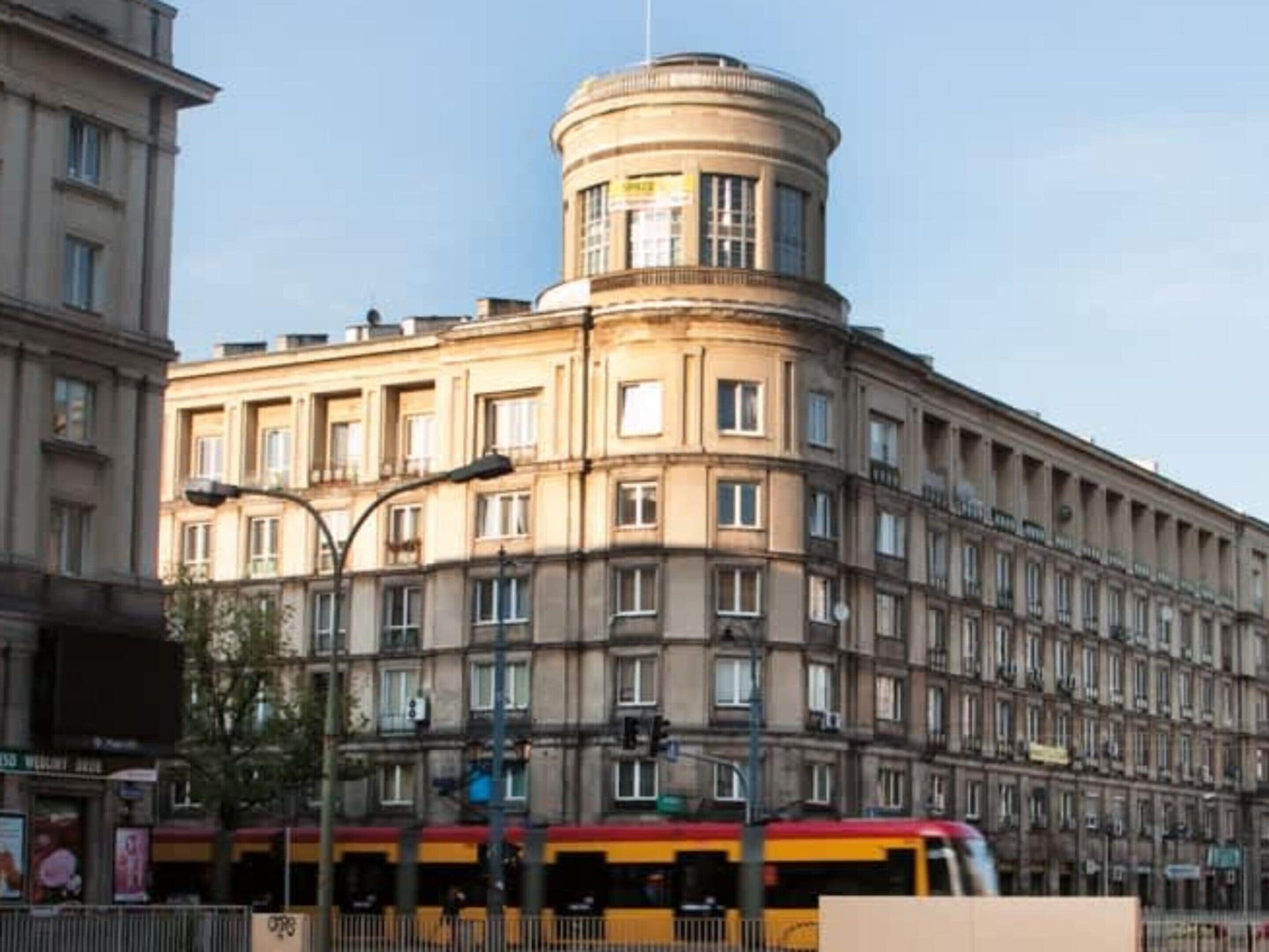Ein mehrstöckiges Gebäude mit einem zentralen Turm, einer Straßenbahnlinie davor und einer Straße mit einer Ampel.