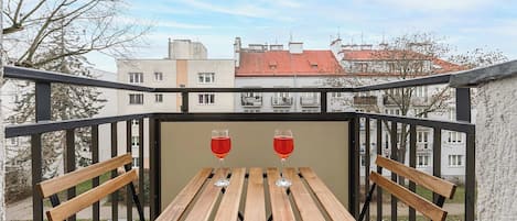 Uma varanda com uma mesa de madeira e duas taças de vinho, com vista para uma área residencial com edifícios e carros estacionados.