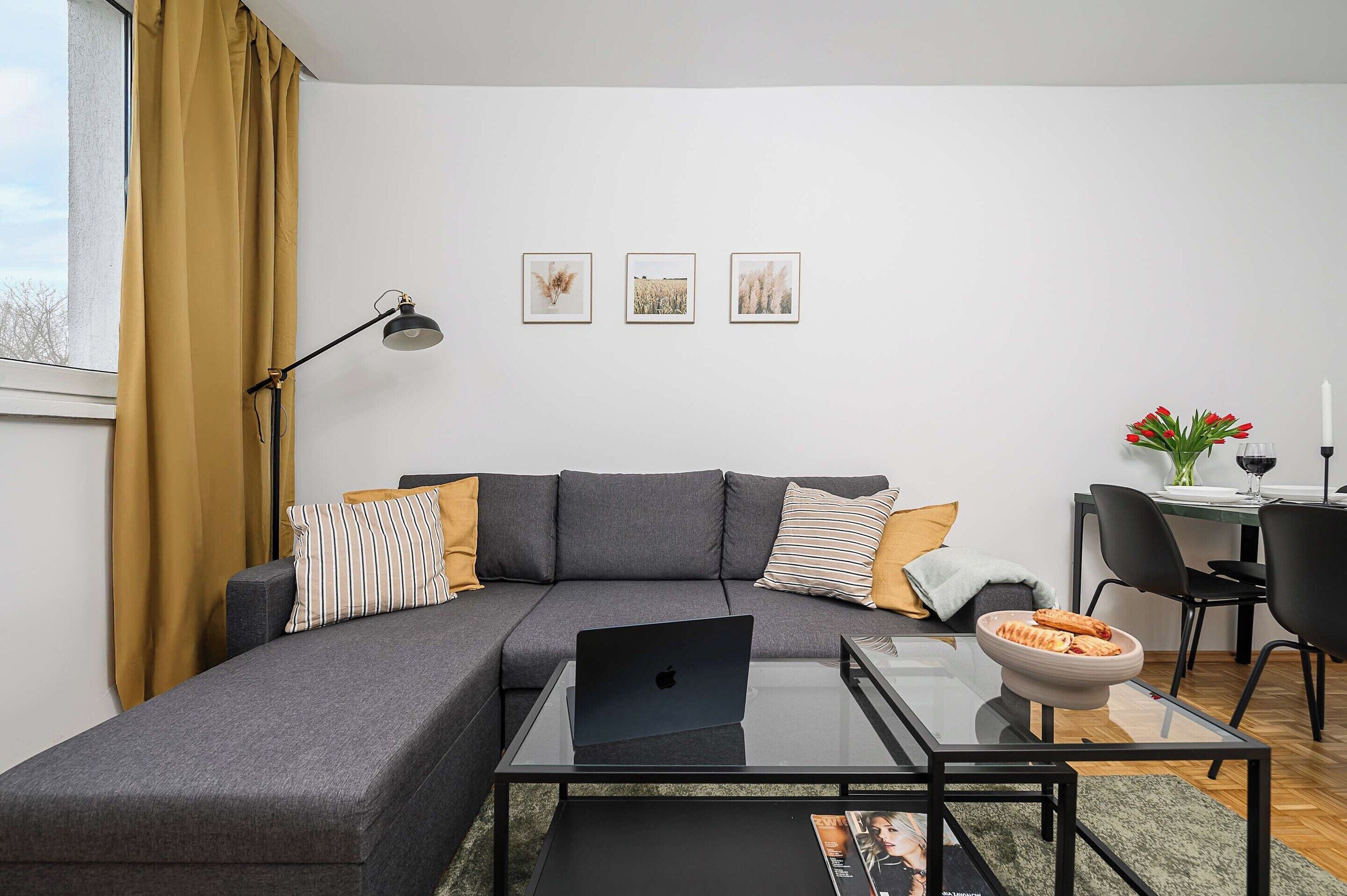 A modern living room with a gray sectional sofa, a glass coffee table, and a laptop. There are three framed pictures on the wall, a desk with a chair, and a dining table with a vase of flowers.