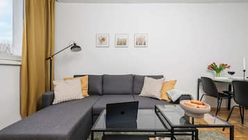Ein modernes Wohnzimmer mit einem grauen Ecksofa, einem Glastisch und einem Laptop. An der Wand hängen drei gerahmte Bilder, ein Schreibtisch mit Stuhl und ein Esstisch mit einer Blumenvase.
