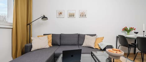A modern living room with a gray sectional sofa, a glass coffee table, and a laptop. There are three framed pictures on the wall, a desk with a chair, and a dining table with a vase of flowers.