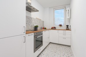 Cocina compacta con armarios blancos, horno de acero inoxidable, encimera de madera y ventana con vistas a árboles desnudos.