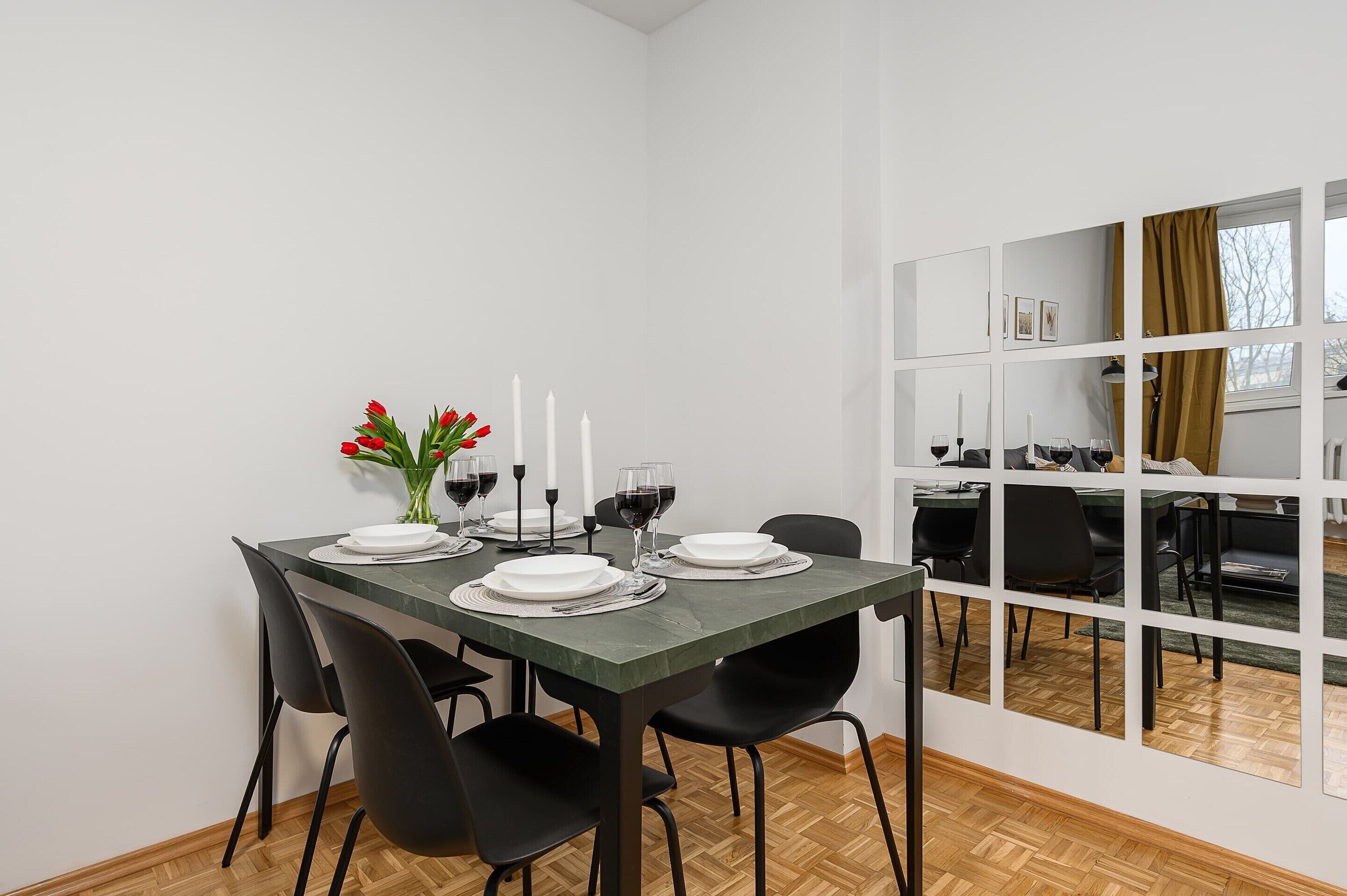 Un coin repas avec une table dressée pour quatre personnes, des chaises, un vase rempli de fleurs rouges et une fenêtre donnant sur un beau paysage.