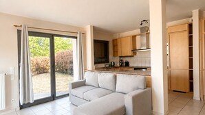 Una sala de estar moderna con un sofá seccional, una zona de cocina con armarios de madera y un gran ventanal corredizo que da a un jardín.