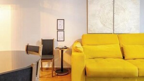 Une pièce avec un canapé jaune, une petite table ronde et une chaise. Deux tableaux encadrés sont accrochés au mur.