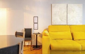 Ein Zimmer mit einem gelben Sofa, einem kleinen runden Tisch und einem Sessel. An der Wand hängen zwei gerahmte Bilder.