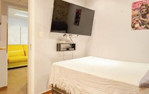 Une chambre à coucher avec un lit, un téléviseur fixé au mur et un canapé jaune dans la pièce adjacente.