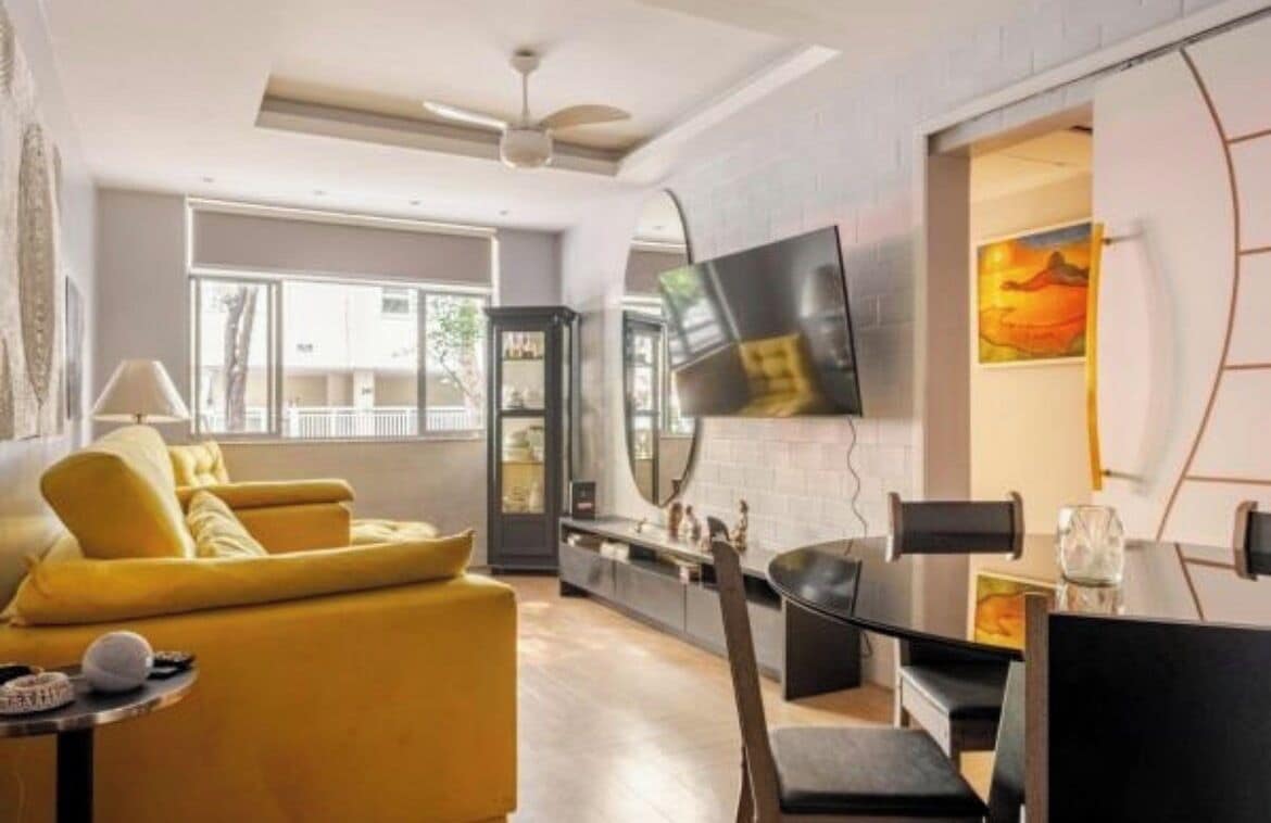 A modern living room with a sofa, a dining table with chairs, a flat-screen TV mounted on the wall, and a large window.