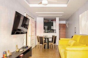 A living room with a yellow sofa, a flat-screen TV mounted on the wall, a dining area with a table and chairs, and a kitchen area visible through an open door. - Modern Copacabana - Ipanema in the best location in Rio de Janeiro. (Rio de Janeiro)