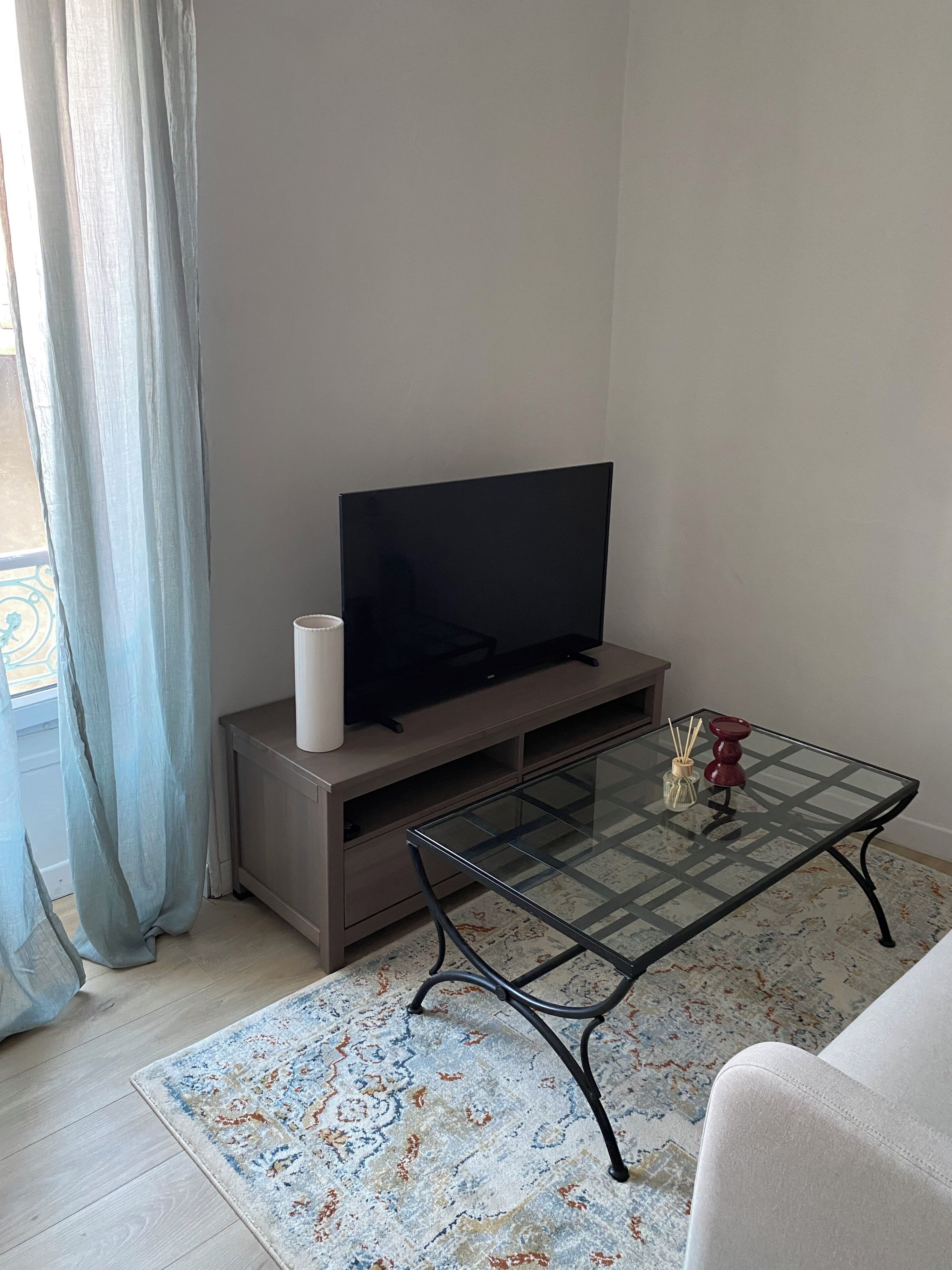 A living room with a flat-screen TV on a stand, a glass coffee table with decorative items, a patterned rug, and a light-colored sofa.