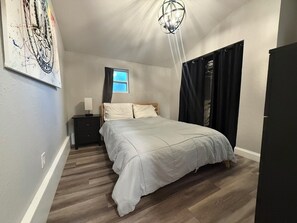 Une chambre à coucher avec un lit, une table de chevet, une lampe, une fenêtre et des rideaux.