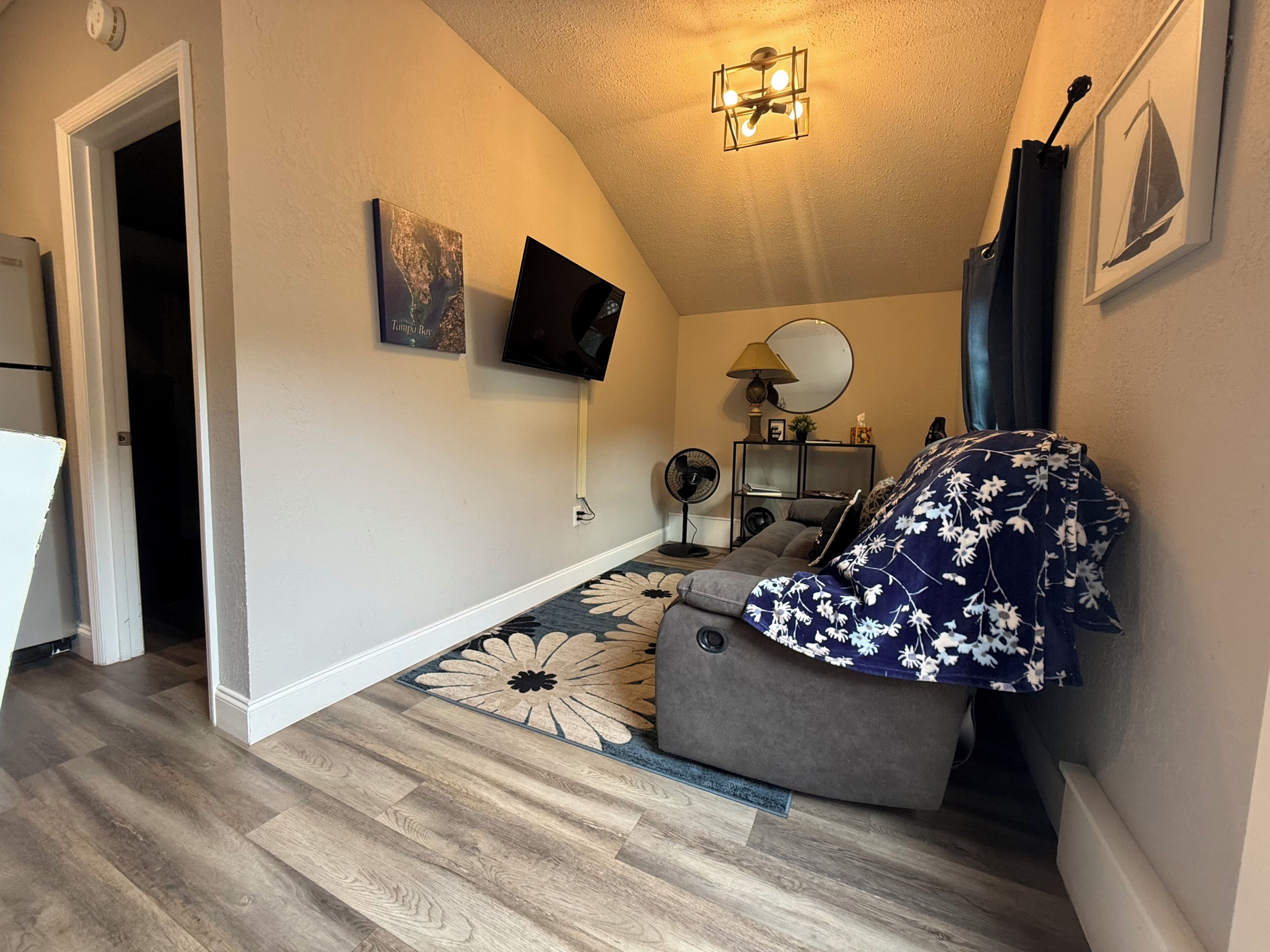 Un espace de vie compact avec un canapé gris, un téléviseur à écran plat fixé au mur, un ventilateur de plafond et un tapis à motifs.
