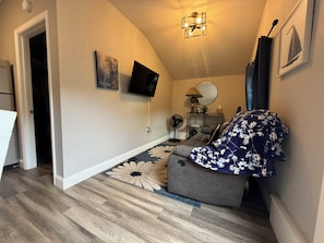 Un espace de vie compact avec un canapé gris, un téléviseur à écran plat fixé au mur, un ventilateur de plafond et un tapis à motifs.