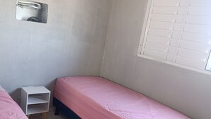 A small, single-bed room with a pink bedspread, a white nightstand, and a window with blinds.
