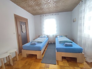 A room with two single beds, a small table, a door, and a window with curtains.