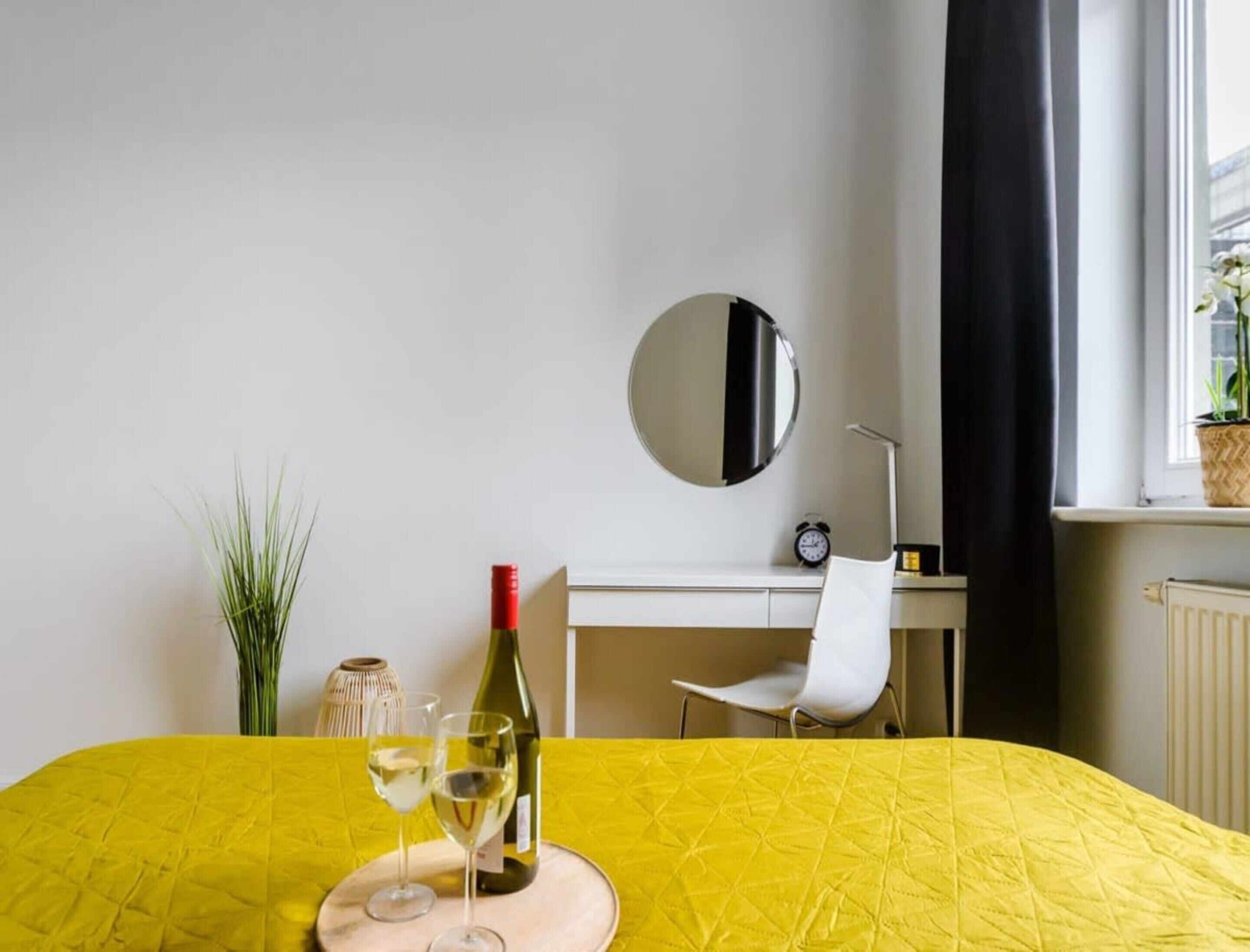 A hotel room with a bed, a desk, a chair, a mirror, a window with curtains, and a potted plant.