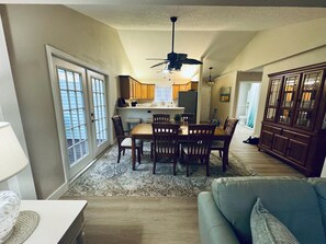 A spacious living area with a dining table, chairs, and a sofa. There is a kitchen in the background with wooden cabinets and a ceiling fan.