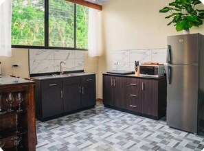 A kitchen with dark cabinets, a stainless steel refrigerator, a microwave, and a window with a view of greenery. - Cozy 1-bedroom house La Fortuna with AC volcano view, swimming pool and Jacuzzi (La Fortuna)