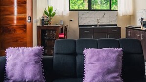 Un salon avec un canapé, deux coussins violets, une porte en bois, une table à manger avec des chaises, une plante et un coin cuisine avec des placards et une fenêtre.