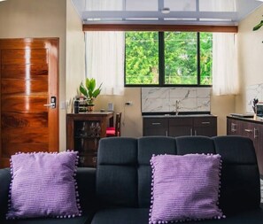 A living room with a sofa, two purple pillows, a wooden door, a dining table with chairs, a plant, and a kitchen area with cabinets and a window. - Cozy 1-bedroom house La Fortuna with AC volcano view, swimming pool and Jacuzzi (La Fortuna)
