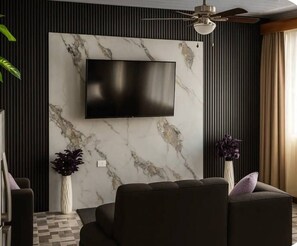 A modern living room with a flat-screen TV mounted on a marble wall, a ceiling fan, and two vases with plants. - Cozy 1-bedroom house La Fortuna with AC volcano view, swimming pool and Jacuzzi (La Fortuna)