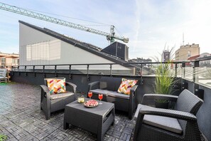 A rooftop terrace with wicker furniture, a table with a watermelon and drinks, and a large building in the background. - Warsaw Flat Near Praga Park (Warsaw)