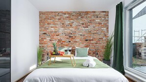 Ein Schlafzimmer mit Backsteinwand, einem Bett mit weißer Bettwäsche, einem Nachttisch aus Holz und einem Fenster mit Blick nach draußen.
