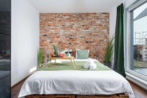 Ein Schlafzimmer mit Backsteinwand, einem Bett mit weißer Bettwäsche, einem Nachttisch aus Holz und einem Fenster mit Blick nach draußen.