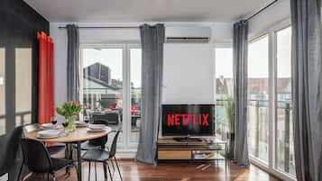 Una sala de estar moderna con comedor, un ventanal con cortinas, un televisor de pantalla plana mostrando 'NETFLIX' y vista a edificios.